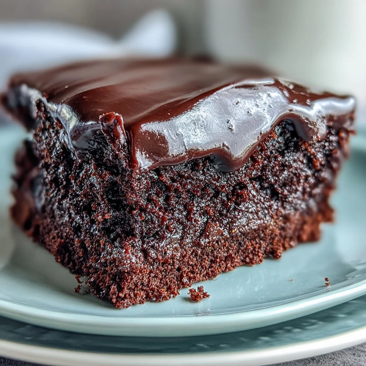 Two layers of moist Easy Chocolate Fudge Cake are covered in rich, glossy chocolate icing on a white plate.