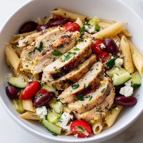 Sliced lemon-garlic chicken rests atop a Greek Chicken Pasta Bowl, with juicy tomatoes, crisp cucumber, and crumbled feta.